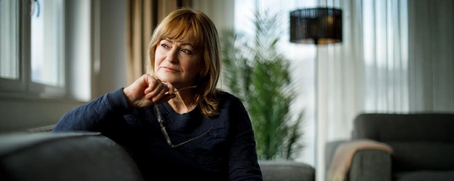 Older woman sitting on a sofa by a window, holding her eyeglasses and looking thoughtfully to the side.