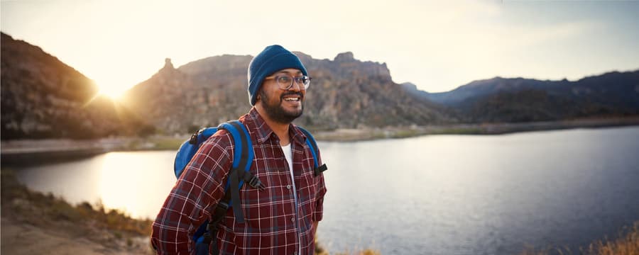 Smiling hiker wearing a blue beanie and backpack standing beside a mountain lake at sunset.