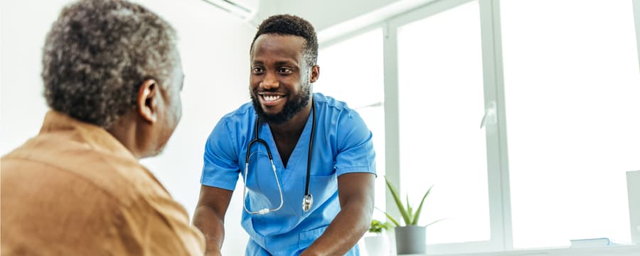 Smiling healthcare worker in blue scrubs and a stethoscope talking with an older man in a bright clinical setting.