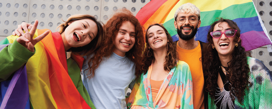 Five smiling people pose together outdoors in front of a large rainbow pride flag. One person is wrapped in a rainbow flag, and the group wears colorful clothing while looking at the camera and celebrating.
