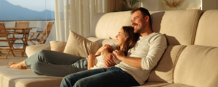A smiling couple relaxes on a beige couch in warm sunlight, with the woman leaning against the man as he wraps his arm around her, both looking content and comfortable in a cozy living room setting.