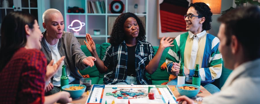 Five adults gathered around a table, laughing and socializing while playing a colorful board game in a cozy living room.