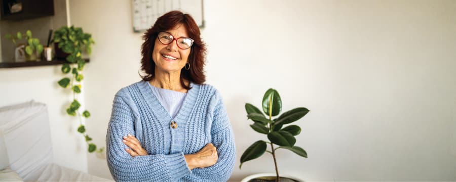 Smiling older woman wearing glasses and a light blue cardigan, standing with her arms crossed in a bright room with houseplants.