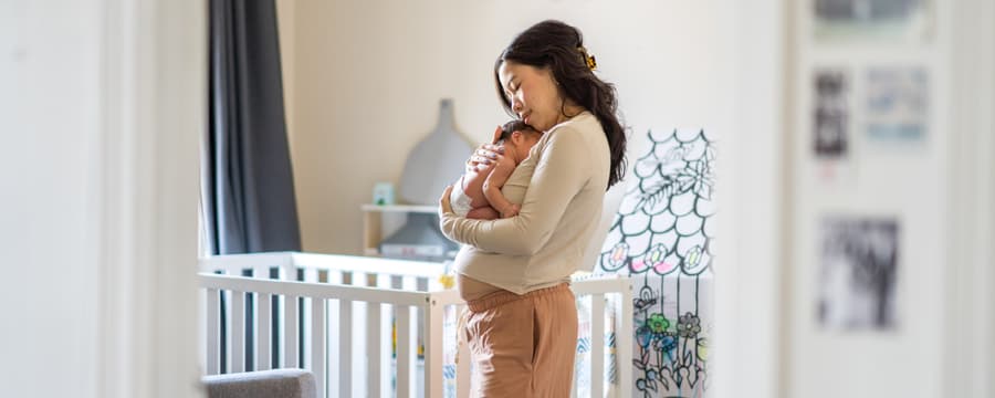 Mother lovingly cradling her newborn baby beside a crib in a softly lit nursery.