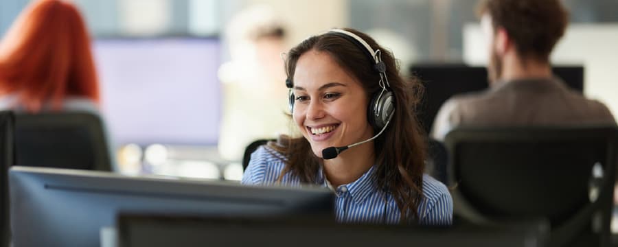 Smiling customer service representative wearing a headset and speaking with a customer in a busy call center.