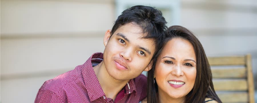 A smiling woman and a teenage boy sit close together outdoors, leaning their heads together and looking at the camera, with a wooden bench and house siding in the background.