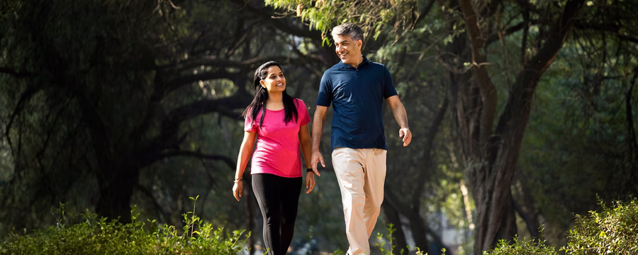 A smiling man and woman walking together on a sunlit path surrounded by trees and greenery.