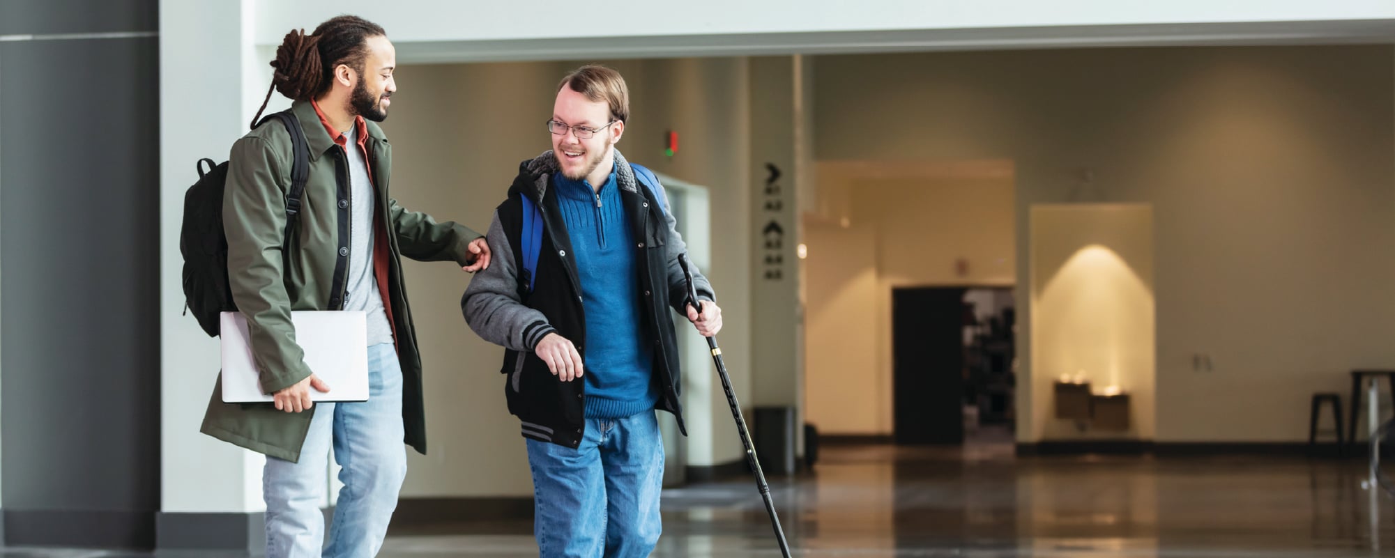 Two people walk together through a spacious indoor building, smiling as they talk. One person carries a white laptop and backpack, while the other uses a white cane and wears a backpack, highlighting an accessible and supportive environment.