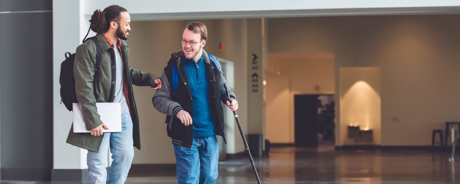 Two people walk together through a spacious indoor building, smiling as they talk. One person carries a white laptop and backpack, while the other uses a white cane and wears a backpack, highlighting an accessible and supportive environment.