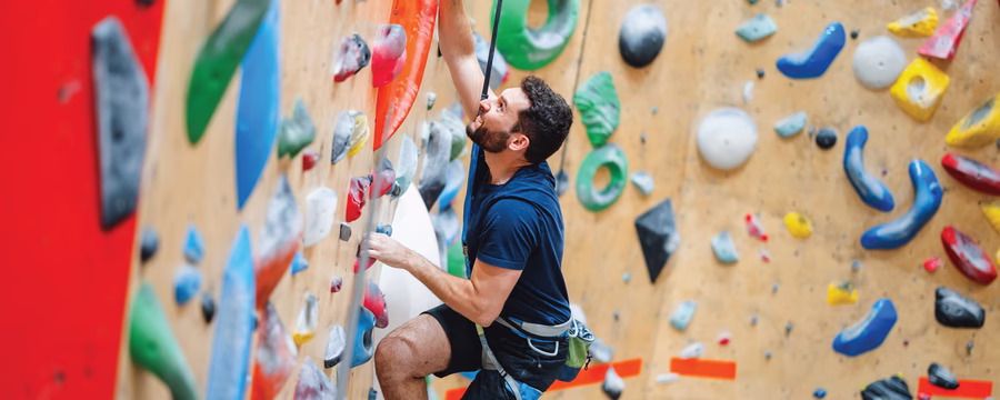 A person wearing a climbing harness and blue athletic clothing climbs an indoor rock wall covered with colorful handholds, secured by a rope in a climbing gym.