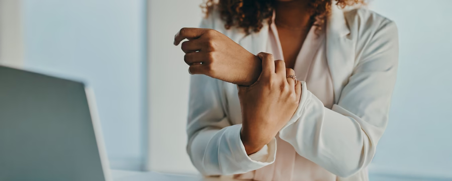 Woman seated at a desk holds her wrist while working on a laptop, suggesting wrist or hand discomfort.