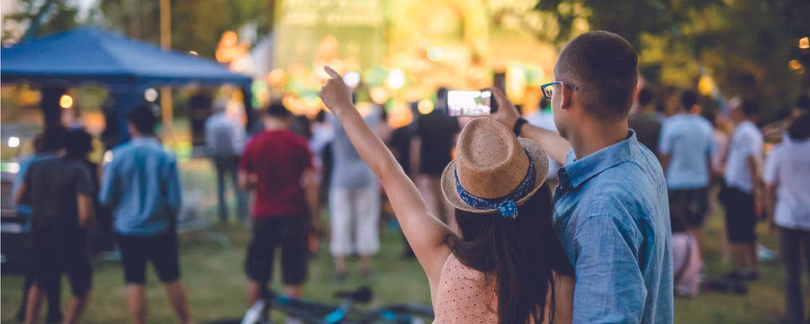 A couple stands arm in arm at an outdoor concert, taking a selfie with a brightly lit stage in the background while a crowd gathers on the grass around them.
