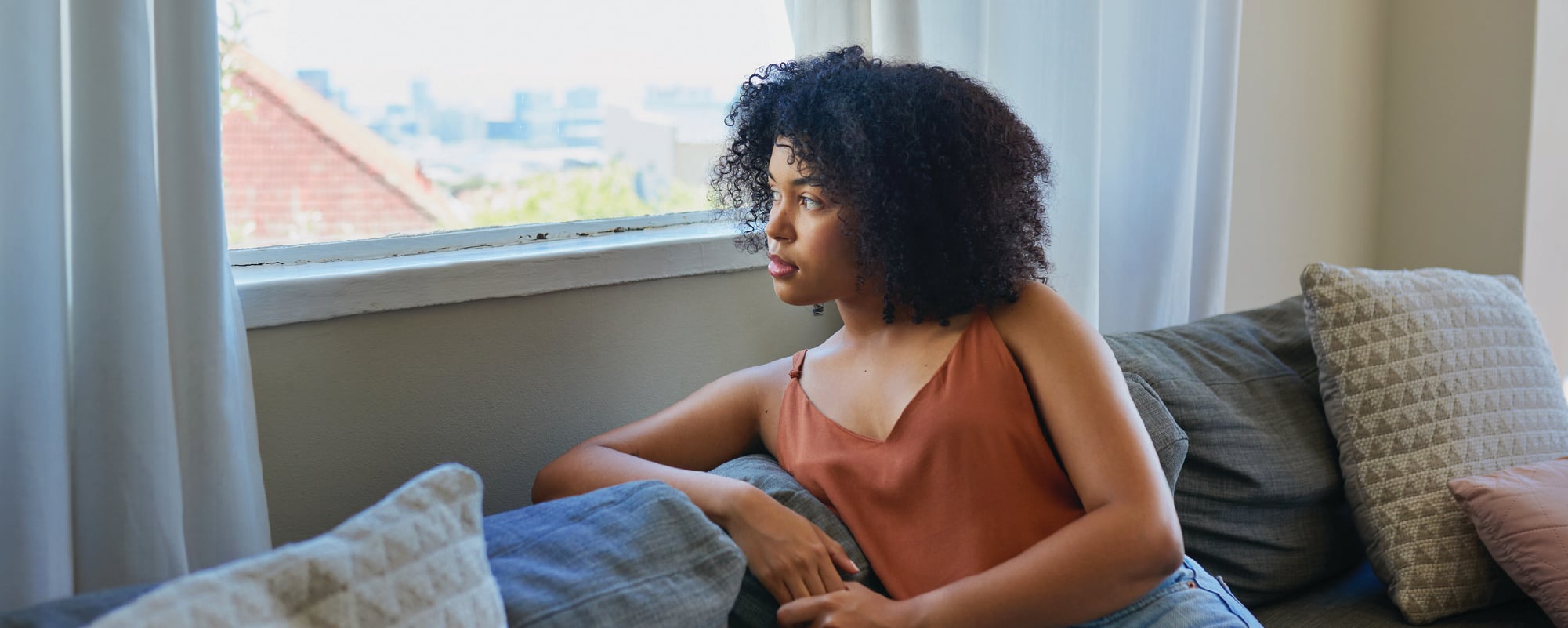 A person with curly hair sits on a couch beside a bright window, gazing thoughtfully outside. Soft natural light fills the living room, creating a quiet, reflective atmosphere with neutral-toned pillows and curtains in the background.