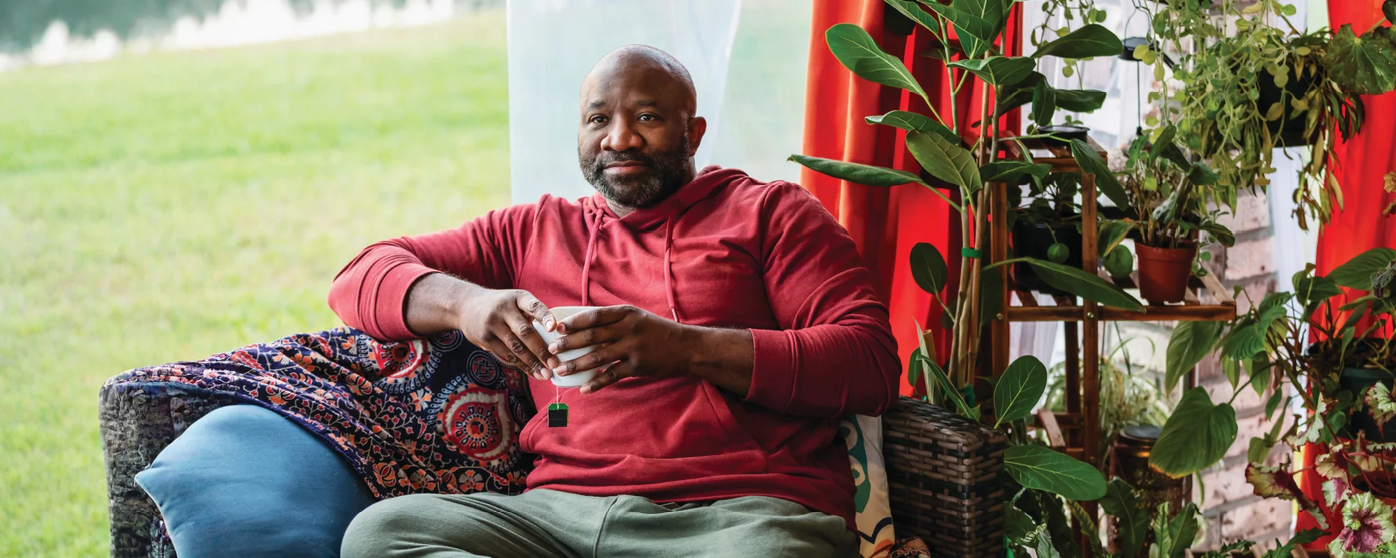 A man in a red hoodie sits comfortably on a sofa holding a mug, surrounded by leafy houseplants beside a bright window.