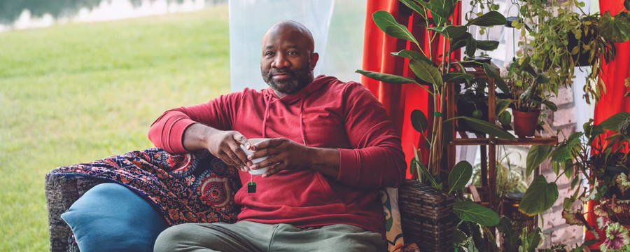 A man in a red hoodie sits comfortably on a sofa holding a mug, surrounded by leafy houseplants beside a bright window.