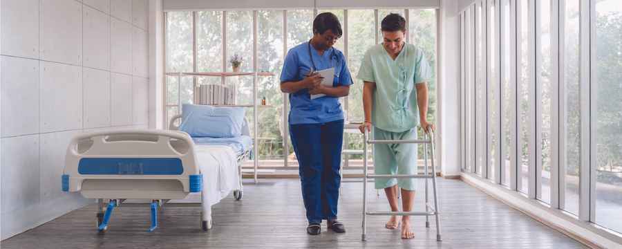 Healthcare professional walks beside and assists a barefoot patient using a walker in a bright hospital rehabilitation hallway.