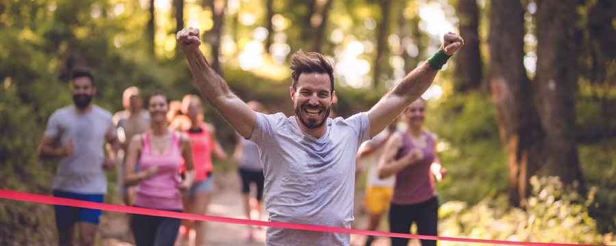 Smiling runner raises his arms in celebration as he crosses the finish line during a race on a wooded trail.