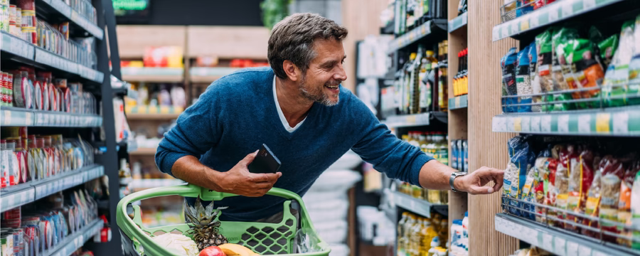 Smiling man shopping for groceries, reaching for an item on a store shelf beside a cart filled with fresh food.
