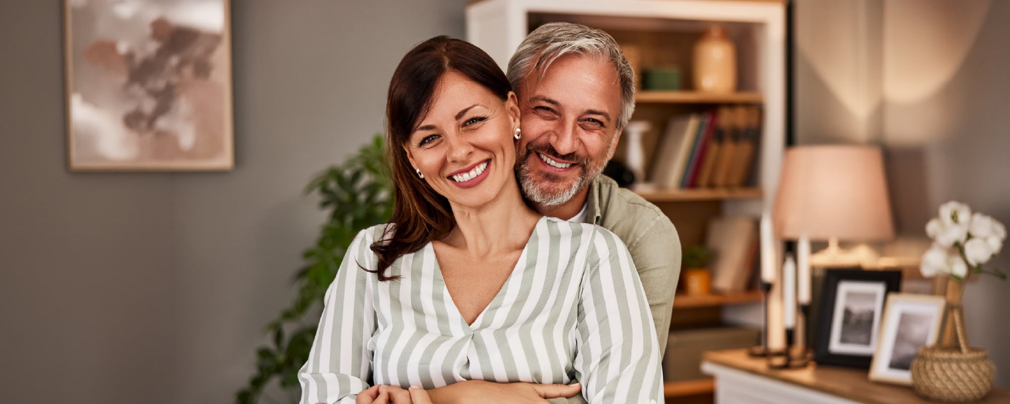 Smiling middle-aged couple embracing in a cozy, modern living room, with the man hugging the woman from behind as both look at the camera. A bookshelf, houseplant, and warm home décor create a welcoming background.