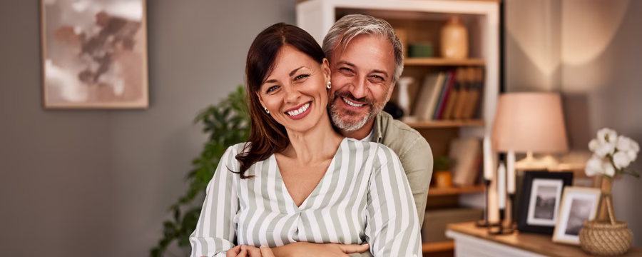 Smiling middle-aged couple embracing in a cozy, modern living room, with the man hugging the woman from behind as both look at the camera. A bookshelf, houseplant, and warm home décor create a welcoming background.