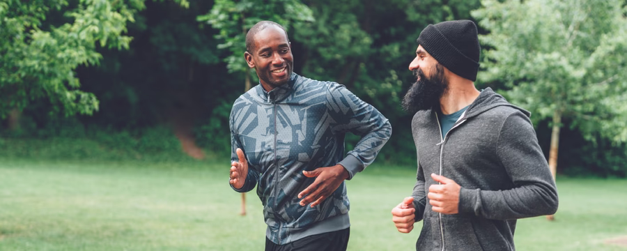 Two adults jogging side by side on a paved path in a green park, smiling and talking as they run. One wears a patterned gray athletic jacket and black leggings, while the other wears a dark beanie, gray hoodie, and blue shorts. Trees and grass fill the background.