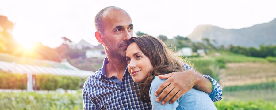 Middle-aged couple embracing outdoors, with the woman resting her head on the man's shoulder as he holds her close.