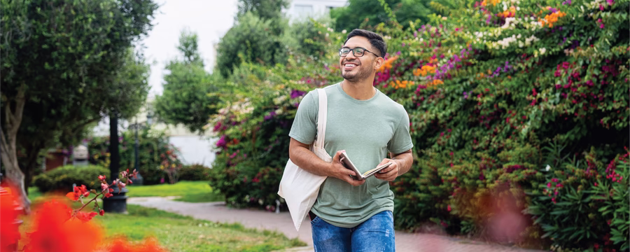 A smiling person wearing glasses, a light green T-shirt, and blue jeans walks along a garden path while holding a notebook. They carry a white tote bag over one shoulder and are surrounded by lush greenery and colorful flowering shrubs. Bright red flowers frame the foreground, and a pigeon stands on the path in the lower right corner.