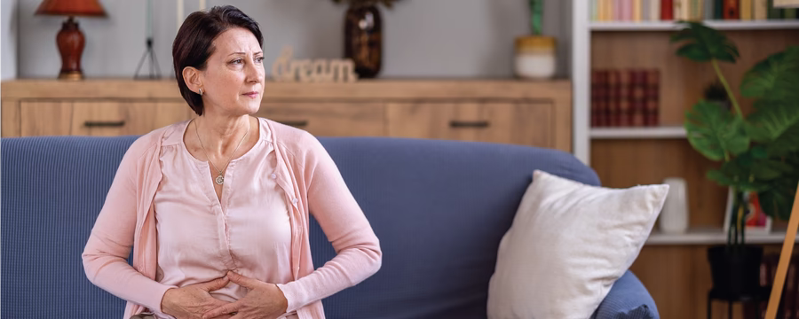 Middle-aged woman sitting on a blue sofa in a living room, holding her lower abdomen with both hands and looking off to the side, appearing uncomfortable or in pain. A lamp, bookshelf, and decorative items are visible in the background.