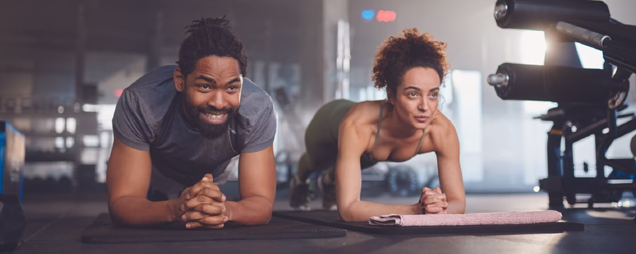 A man and woman hold plank positions on exercise mats while working out together in a gym.