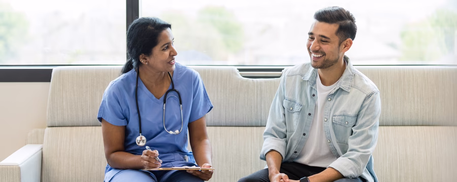 Smiling male patient talking with a female healthcare provider in scrubs, discussing health in a relaxed clinical setting