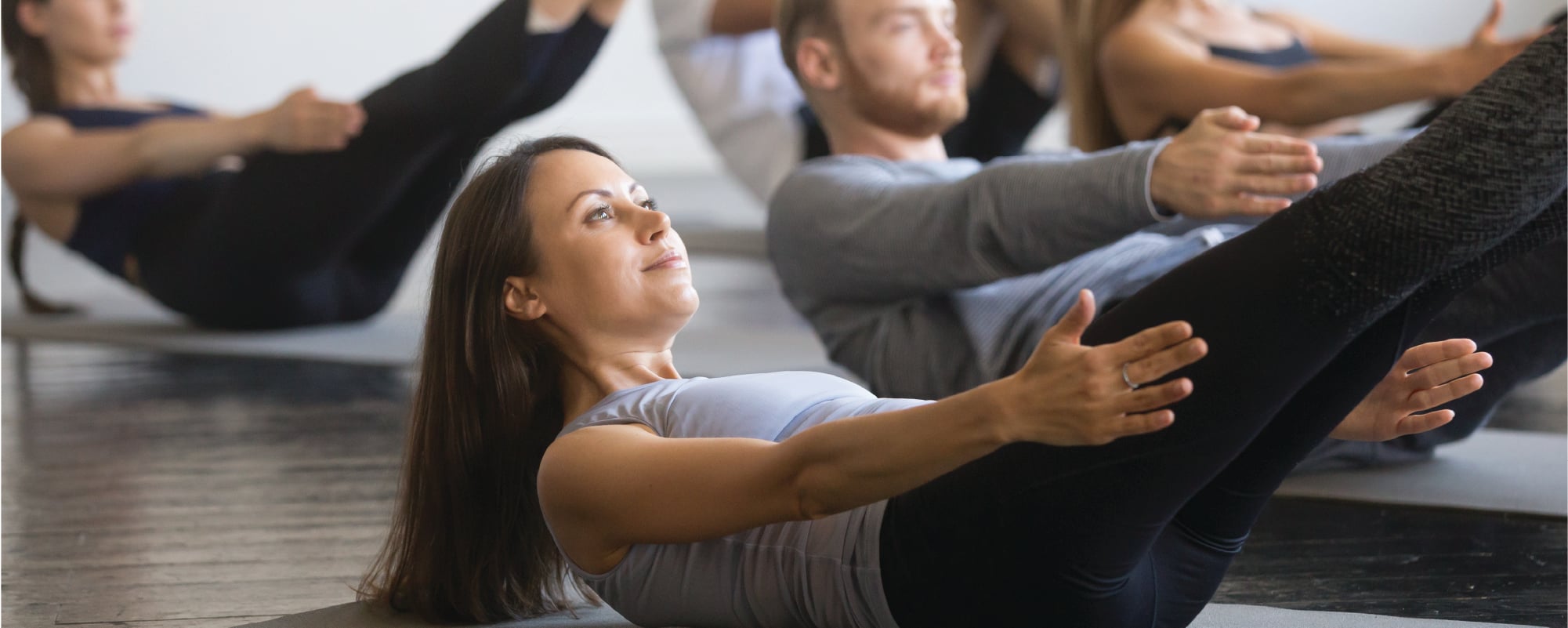 A group fitness class performs core-strengthening exercises on yoga mats, with participants holding a V-sit position with arms extended and legs raised in a bright exercise studio.