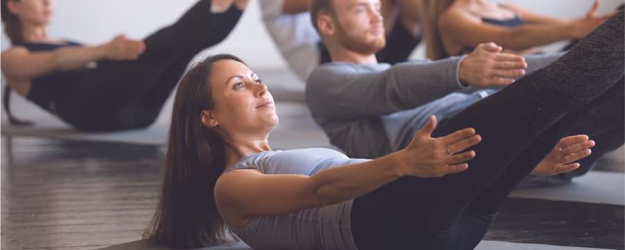 A group fitness class performs core-strengthening exercises on yoga mats, with participants holding a V-sit position with arms extended and legs raised in a bright exercise studio.