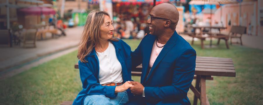 Two people sitting closely together outdoors, smiling and holding hands while talking. One person wears a blue blazer and glasses, and the other wears a blue jacket over a white shirt. A blurred park or market scene appears in the background.