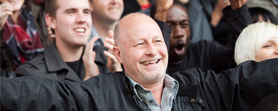 A bald man in a black jacket and light blue shirt stands at the center of a smiling group of diverse people, his arms raised in celebration. The crowd behind him appears joyful and supportive, suggesting a moment of community encouragement or shared accomplishment.