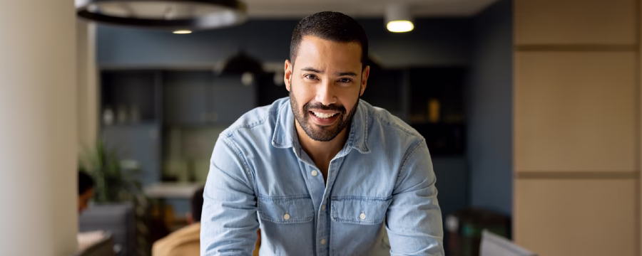 Smiling man in a light blue button-down shirt leans on a kitchen island with folded hands in a modern home, looking directly at the camera.