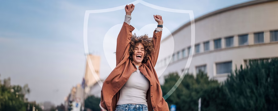 Smiling person outdoors with arms raised in celebration in front of a subtle heart-and-shield graphic, symbolizing confidence and protection.