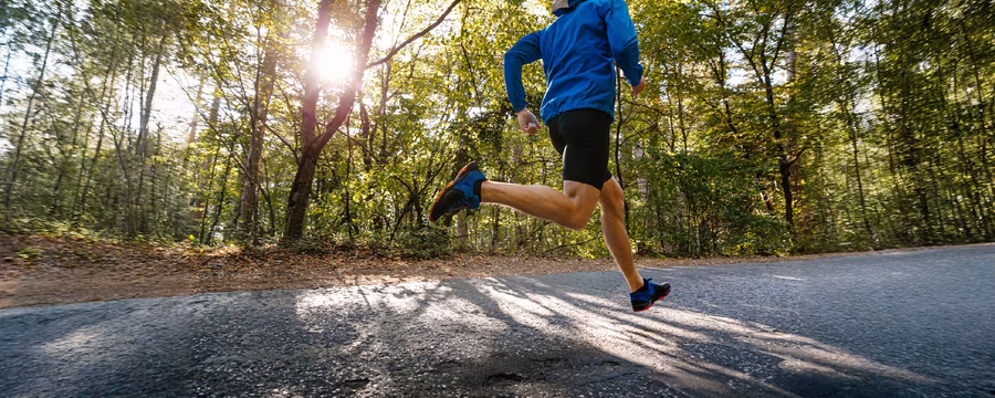 A runner in a blue jacket, black shorts, and blue running shoes sprints along a sunlit paved trail through a wooded park, with sunlight filtering through the trees and casting long shadows across the path.