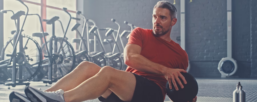 A man in a red athletic shirt and black shorts performs a seated core exercise on a gym mat, twisting his torso while holding a medicine ball. Exercise equipment like stationary bikes is visible in the background, and a towel lies on the floor nearby.