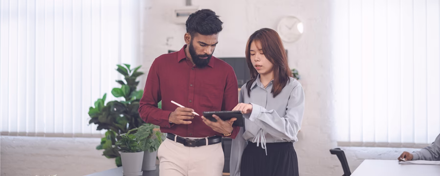 Two coworkers stand in a bright office, reviewing information together on a tablet while one holds a stylus.