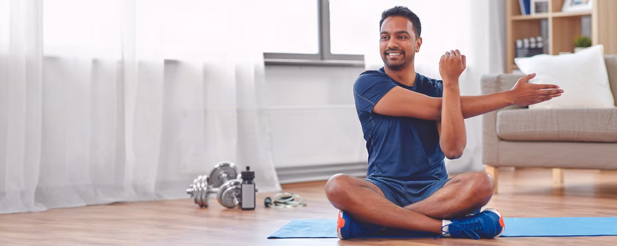 Smiling man sitting cross-legged on an exercise mat and stretching his arm during a home workout.