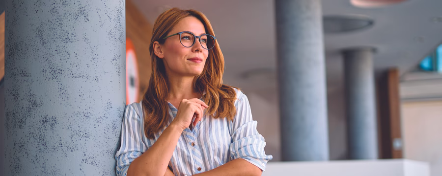 Woman wearing glasses leans against a column in a modern office, looking thoughtfully into the distance.