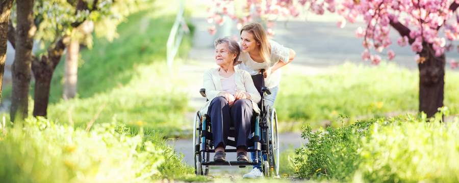 Older woman in a wheelchair enjoying a sunny park with a smiling caregiver standing beside her under blooming pink trees.