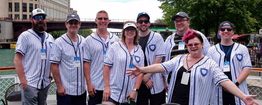 A group of eight adults at NorthShore's Day Out event wearing matching pinstriped baseball jerseys smiles for a photo at a baseball stadium. They stand together near the seating area with the field, scoreboard, and stadium structure visible in the background on a sunny day.