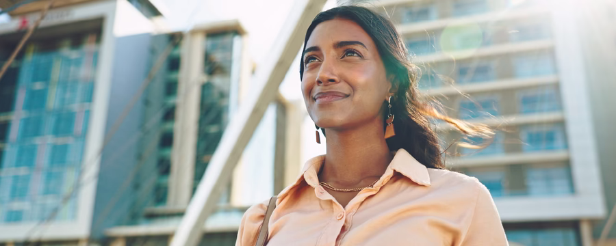 A woman in a light blouse stands outdoors in front of modern buildings, smiling slightly and looking ahead, with sunlight creating a warm glow around her.