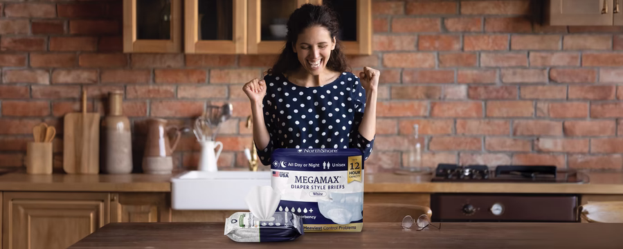 A smiling woman in a cozy kitchen raises her fists in excitement while looking at a package of NorthShore MegaMax diaper-style briefs on the table, with a pack of wipes beside it.