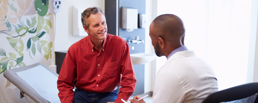A healthcare provider consults with a middle-aged patient seated on an exam table in a medical exam room, listening attentively and taking notes during a face-to-face appointment.