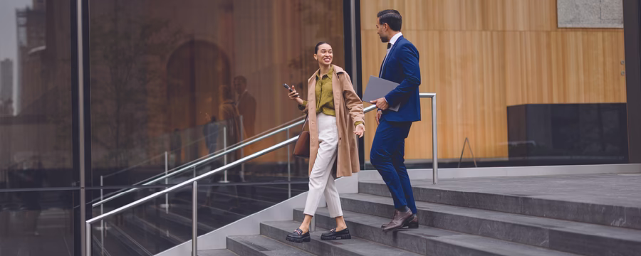Two young professionals talking while walking down outdoor steps of a modern office building, one holding a smartphone and the other carrying a laptop.