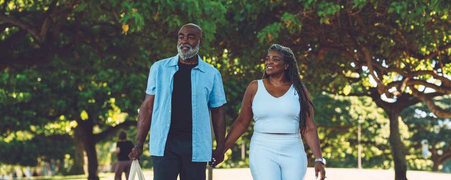Smiling couple holding hands while walking together on a tree-lined path in a sunny park.