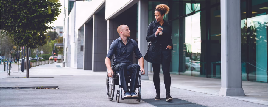 Man using a wheelchair talks with a woman holding a coffee as they walk together outside a modern office building.