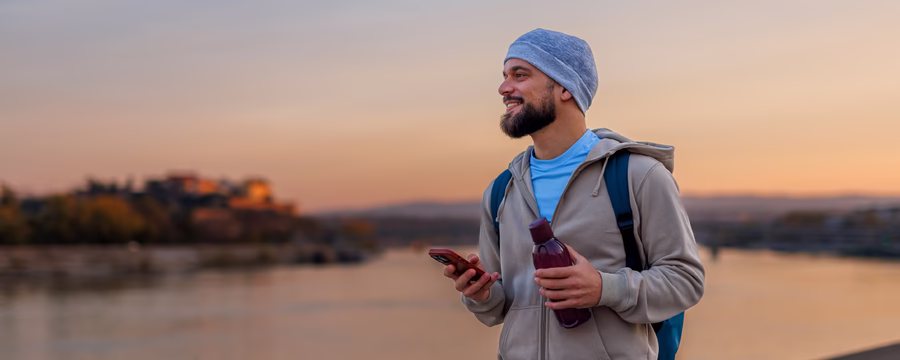 A bearded man wearing a blue beanie and gray jacket stands outdoors at sunset, holding a smartphone and water bottle with a backpack on his shoulders.